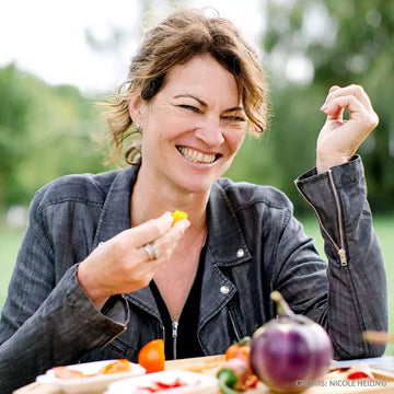Eine Frau lacht, hält Essen in einem Park, mit Gemüse auf einem Tisch.