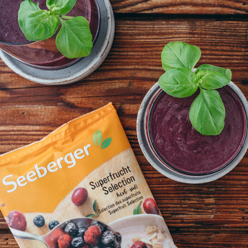 Ein dunkler Smoothie mit Basilikum auf Holztisch, neben Seeberger Superfrucht Selection Packung.