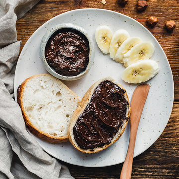 Brot mit Schokoladenaufstrich, Bananenscheiben daneben, auf einem Teller, umgeben von Haselnüssen.