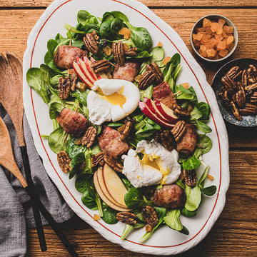 Salat mit pochierten Eiern, Speck, Apfelstücken und gerösteten Nüssen auf weißem Teller angerichtet.