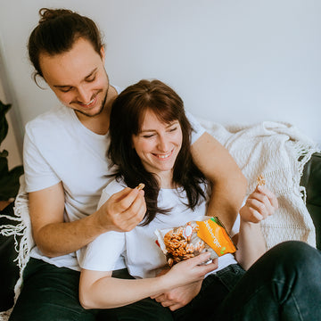 Pärchen sitzt lachend auf Sofa, isst Snacks aus einer offenen Tüte.